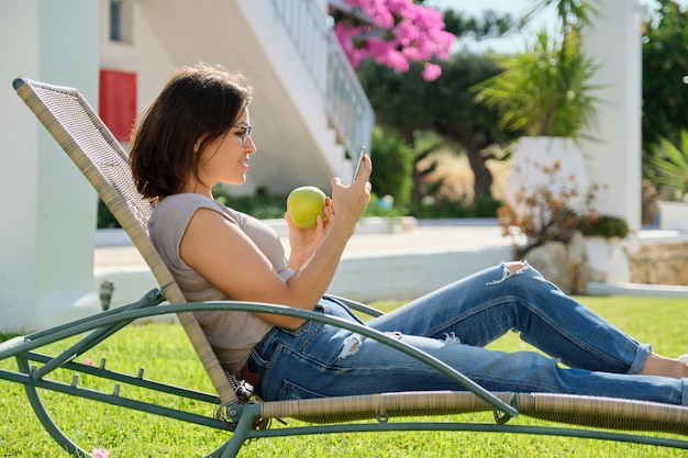 Hermosa mujer madura descansando en una hamaca sobre césped verde. Mujer relajante comiendo manzana y usando smartphone, día soleado de verano. Estilo de vida, ocio, nutrición, personas de mediana edad.