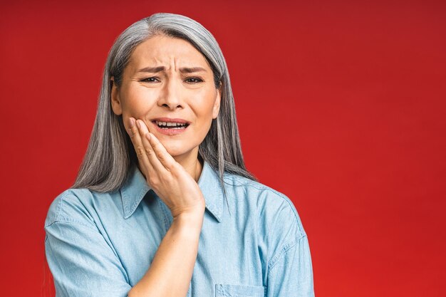 Hermosa mujer madura de edad avanzada asiática tocando la boca con la mano con expresión dolorosa debido a dolor de muelas o enfermedad dental en los dientes Concepto de dentista aislado sobre fondo rojo