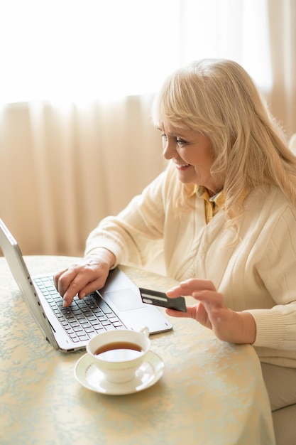 Hermosa mujer madura realiza compras en Internet con una tarjeta de crédito mientras está sentada a la mesa y toma una taza de té