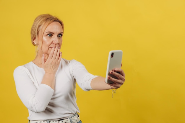 Hermosa mujer madura con un teléfono en la mano sorprendida por lo que vio en la pantalla con un fondo amarillo