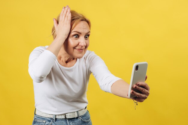 Hermosa mujer madura con un teléfono en la mano sorprendida por lo que vio en la pantalla con un fondo amarillo