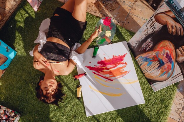 Hermosa mujer con manchas de pintura en su cuerpo yace sobre una alfombra de hierba verde con los ojos cerrados entre paquetes de lienzos de retratos de paletas de pinturas