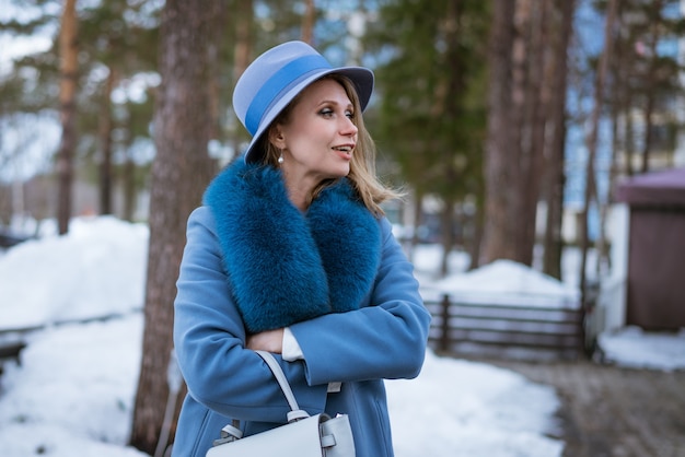 Hermosa mujer con maquillaje, abrigo azul y sombrero, posando en la naturaleza en la nieve.