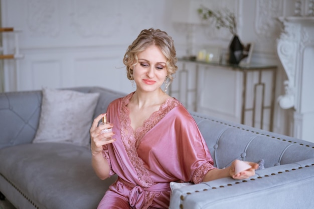 Hermosa mujer con maquillaje y peinado en bata de seda rosa rocía perfume