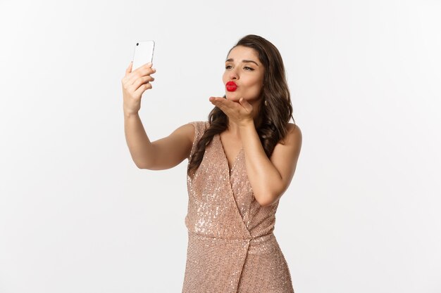 Hermosa mujer con maquillaje y peinado, vestido con glamour, tomando selfie en teléfono inteligente, celebrando la fiesta de año nuevo, fondo blanco