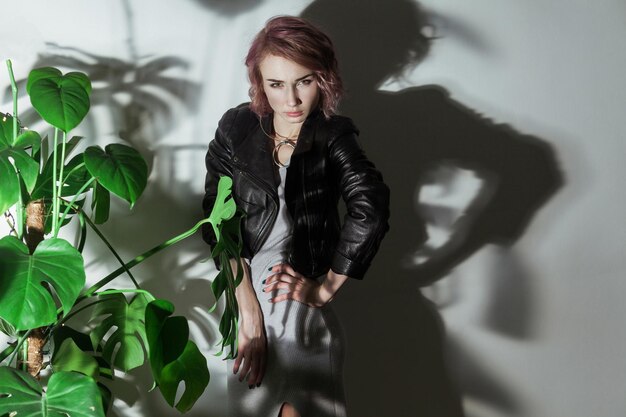 Hermosa mujer con maquillaje y pelo rosa con gris y chaqueta de cuero negro posando cerca de hojas de palma verdes sobre fondo tiro de estudio interior. | Foto