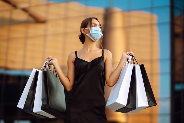 Hermosa mujer en máscara médica protectora estéril con bolsas de compras cerca del centro comercial.