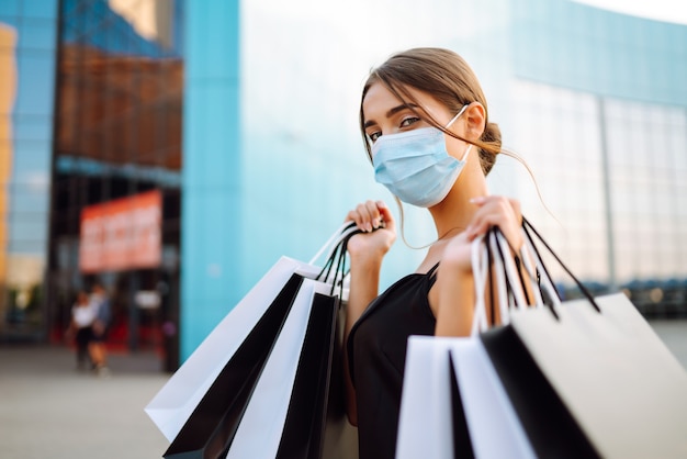 Hermosa mujer en máscara médica protectora estéril con bolsas de compras cerca del centro comercial.