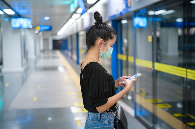 Una hermosa mujer con una mascarilla quirúrgica con teléfono mientras espera un tren en el metro, la tecnología y el concepto covid19