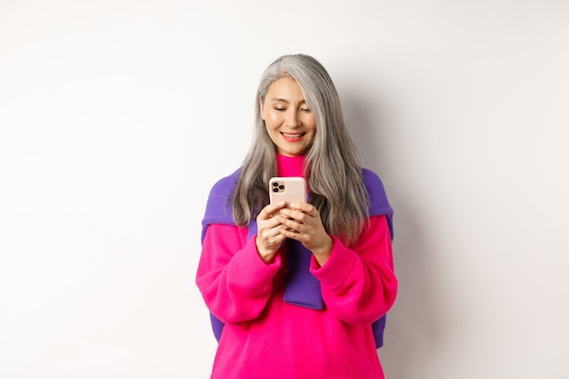 Hermosa mujer mayor asiática en suéter con estilo rosa con smartphone. Anciana leyendo en el teléfono móvil y sonriendo, de pie sobre fondo blanco.