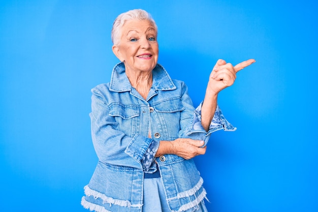 Hermosa mujer mayor con ojos azules y cabello gris con estilo casual de mezclilla sonriendo feliz señalando con la mano y el dedo al lado