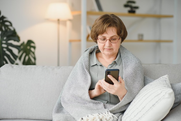 Hermosa mujer mayor en ropa casual usando su teléfono inteligente mientras está sentado en el sofá en casa
