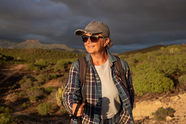 Hermosa mujer mayor con sombrero y ropa deportiva caminando en la naturaleza a la luz del atardecer disfrutando de la libertad y las vacaciones Mujer madura feliz con bastón mirando el paisaje tropical de montaña