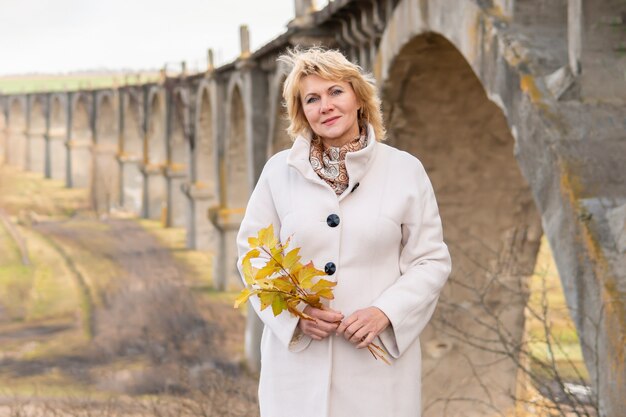 Una hermosa mujer de mediana edad camina en el parque natural en otoño. Ella está de pie junto al puente viejo.