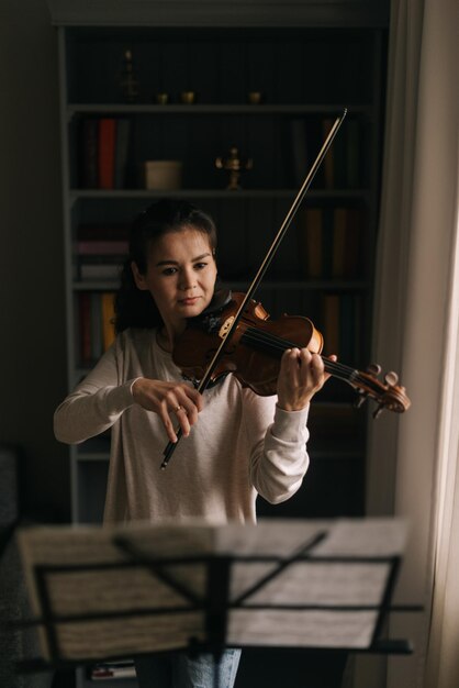 Hermosa mujer mira la partitura mientras toca el violín La niña está practicando tocar un instrumento musical en casa