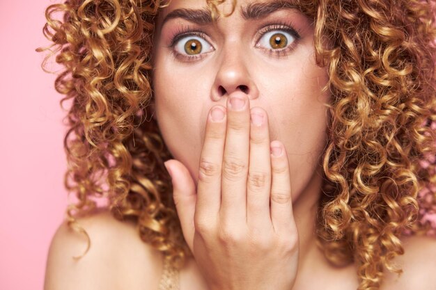 Hermosa mujer mirando a la cámara con sorpresa y tapándose la boca con las manos maquillaje brillante fondo rosa