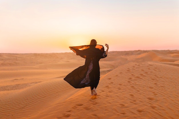 Hermosa mujer misteriosa en vestido largo negro árabe tradicional se encuentra en el desierto al atardecer