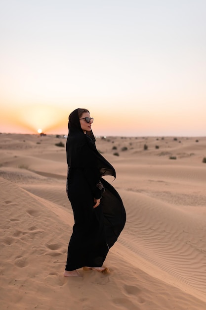 Hermosa mujer misteriosa en vestido largo negro árabe tradicional se encuentra en el desierto al atardecer