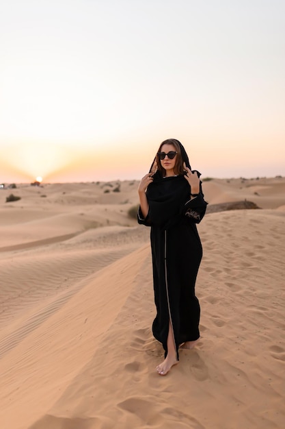 Hermosa mujer misteriosa en vestido largo negro árabe tradicional se encuentra en el desierto al atardecer