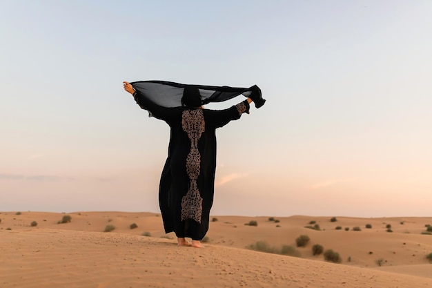 Hermosa mujer misteriosa en vestido largo negro árabe tradicional se encuentra en el desierto al atardecer