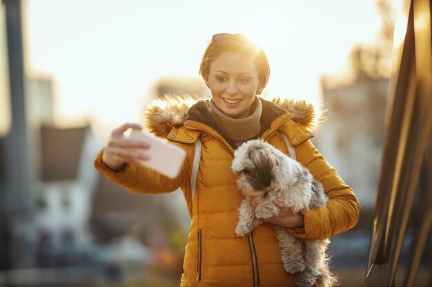 Hermosa mujer de moda joven está pasando tiempo con su lindo perro mascota en la calle de la ciudad y haciendo selfie.