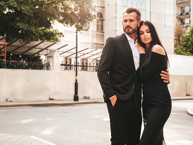 Hermosa mujer de moda y su novio elegante guapo en traje Modelo morena sexy en vestido de noche negro Pareja de moda posando en la calle en Europa Hombre brutal y su mujer al aire libre