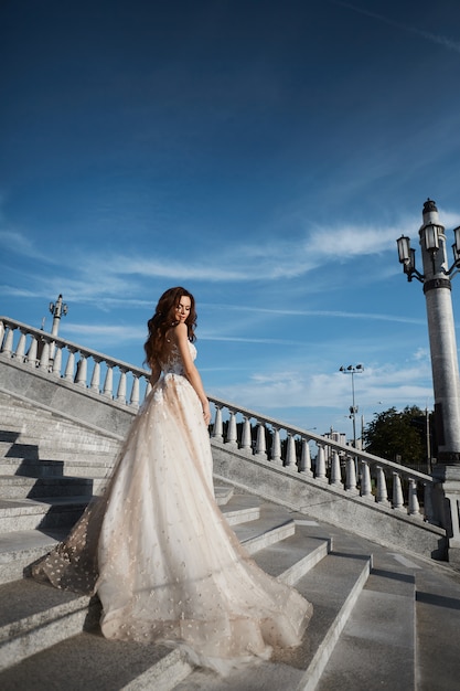 Hermosa mujer modelo con cuerpo perfecto en vestido de novia de lujo se encuentra con la espalda en las escaleras y posando con cielo azul en el fondo