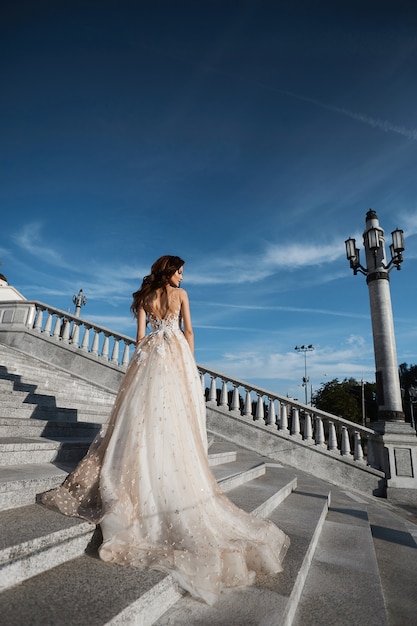 Hermosa mujer modelo con cuerpo perfecto en vestido de novia de lujo se encuentra con la espalda en las escaleras y posando con cielo azul en el fondo