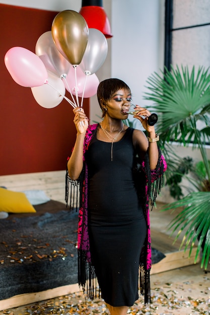 Hermosa mujer modelo negro con globos y una copa de champán preparándose para la fiesta de cumpleaños. Refinada niña africana en vestido negro con sonrisa después del evento.