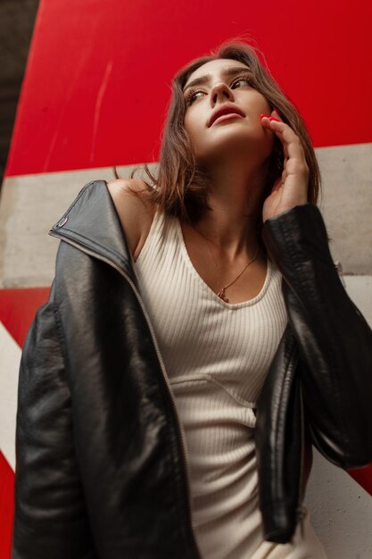 Hermosa mujer modelo sexy de moda en ropa urbana de moda con una chaqueta de cuero y vestido posando en la calle cerca de la pared de hormigón rojo