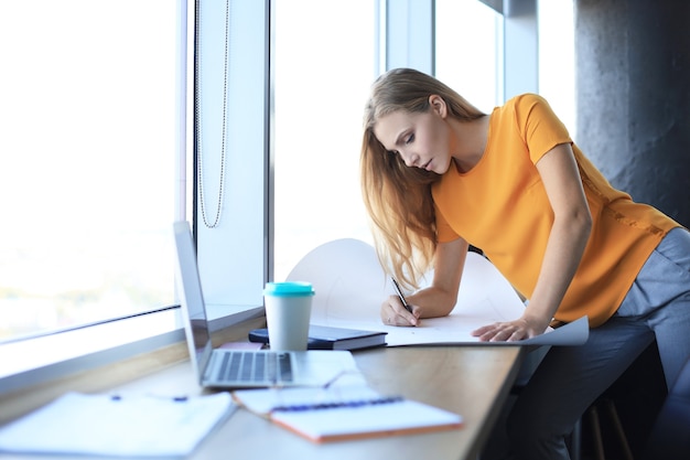 Hermosa mujer moderna está escribiendo algo en el plano mientras trabaja en la oficina creativa.