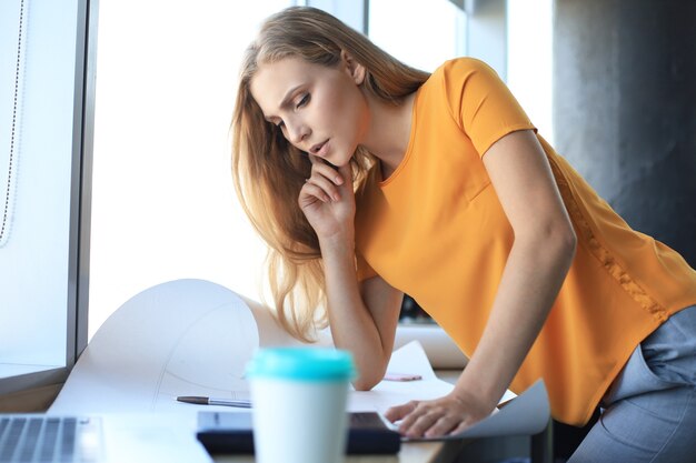 Hermosa mujer moderna está escribiendo algo en el plano mientras trabaja en la oficina creativa.