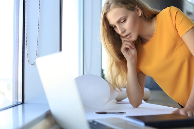 Hermosa mujer moderna está escribiendo algo en el plano mientras trabaja en la oficina creativa.