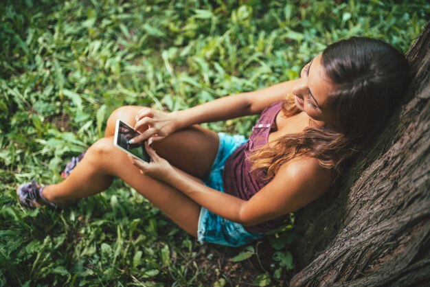 Hermosa mujer moderna sonriente enviando mensajes de texto en un teléfono inteligente sentado debajo de un árbol en un parque de la ciudad