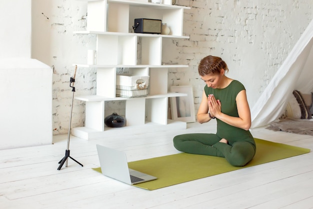 Una hermosa mujer con un mono deportivo verde en una habitación luminosa está transmitiendo en línea realiza un yoga