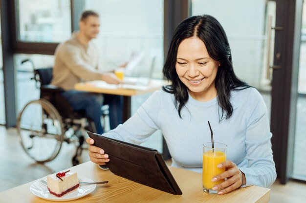 Hermosa mujer morena alerta sonriendo bebiendo jugo y usando su tableta mientras está sentada en un café y un hombre en silla de ruedas sentado en el fondo