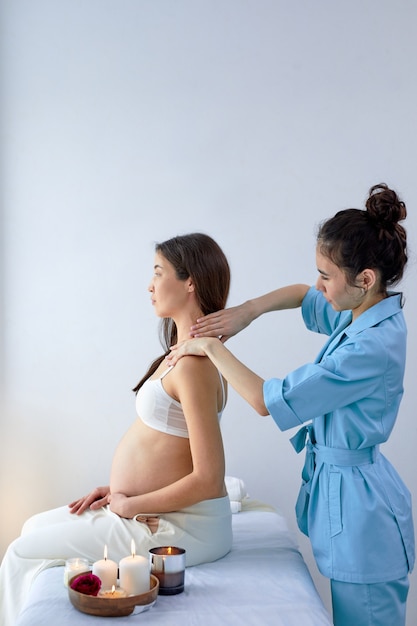 Hermosa mujer morena asiática grávida disfrutando de masaje de espalda y hombros en la sala de esteticista en el centro de spa, vista lateral de la relajada dama sentada en la cama, descansando. fisioterapeuta haciendo masaje