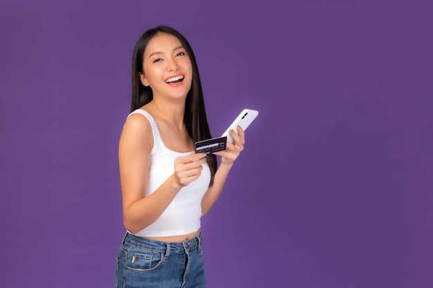 Hermosa mujer morena asiática linda chica en camiseta blanca con tarjeta de crédito y teléfono móvil inteligente utiliza una banca en línea para pagar compras en línea aisladas en un fondo morado