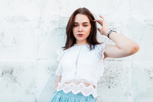 Hermosa mujer morena bastante joven con maquillaje natural en un top de encaje en pantalones azules vintage posa cerca de una pared blanca