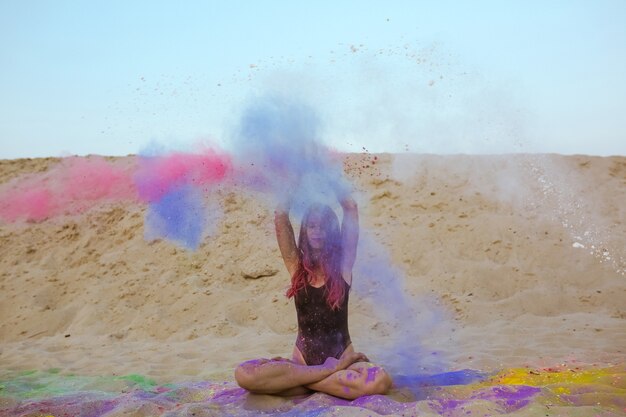 Hermosa mujer morena con cabello largo jugando con pintura Holi, sentada en la arena