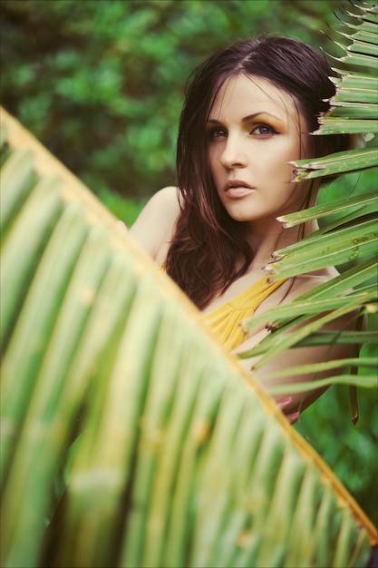Hermosa mujer morena con cabello mojado en vestido largo amarillo rodeado de plantas tropicales