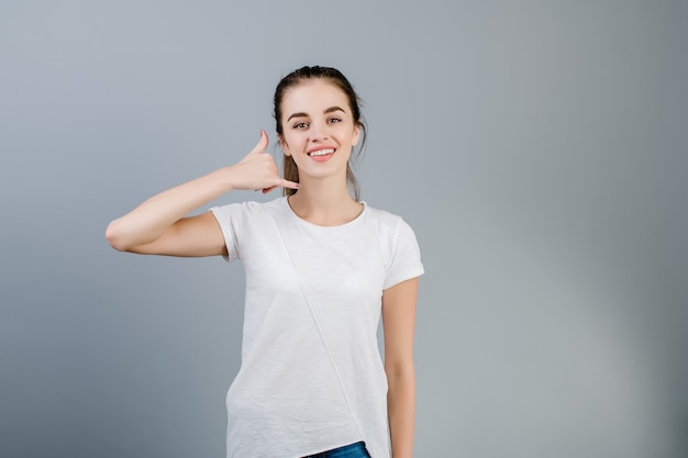 Hermosa mujer morena con camisa blanca mostrando llamarme gesto imitando teléfono con mano aislado sobre gris