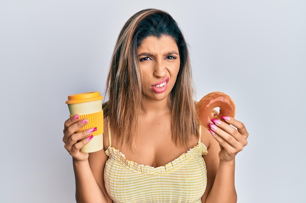 Hermosa mujer morena comiendo donas y bebiendo café concepto de duda de expresión despistada y confusa