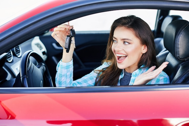 Hermosa mujer morena conduciendo un coche rojo, compra de automóviles. Feliz propietario de coche nuevo, mirando las llaves del coche y sonriendo. Cabeza y hombros, conductor feliz