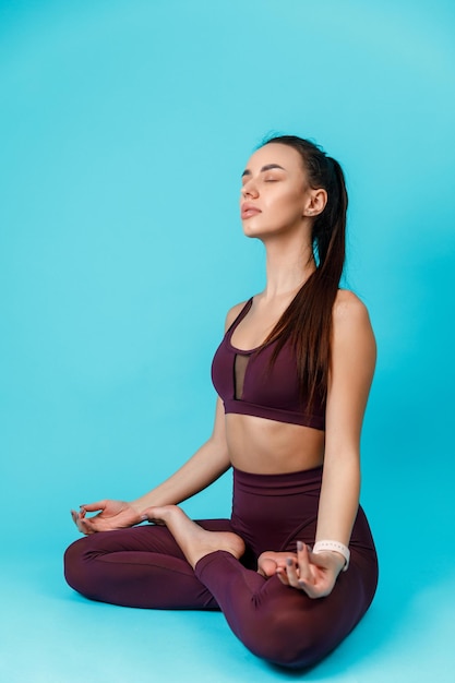 Hermosa mujer morena deportiva se dedica al yoga, se sienta en la posición de loto sobre un fondo azul. ejercicios deportivos aislado