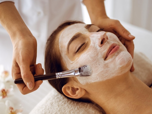 Hermosa mujer morena disfrutando aplicando máscara cosmética con los ojos cerrados. Tratamiento relajante en conceptos de centro de medicina y spa.