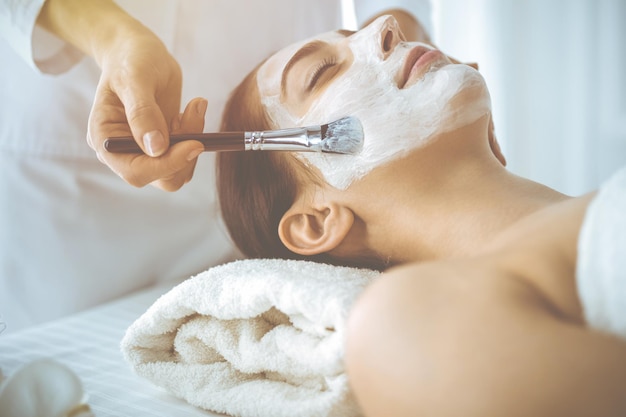 Hermosa mujer morena disfrutando aplicando máscara cosmética con los ojos cerrados. Tratamiento relajante en conceptos de medicina y spa.