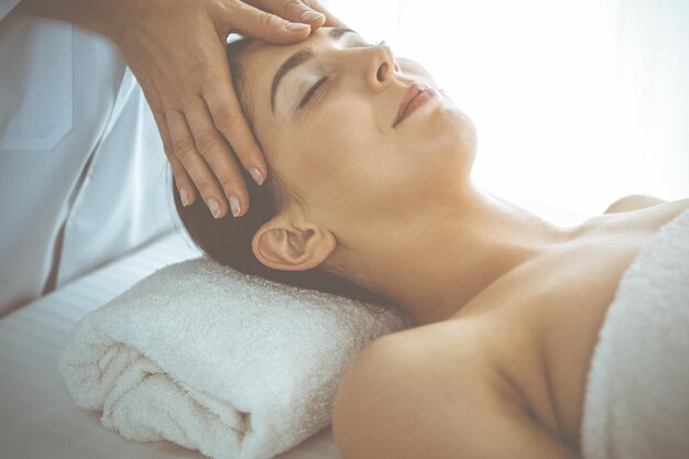 Hermosa mujer morena disfrutando de un masaje facial con los ojos cerrados en el soleado centro de spa. Tratamiento relajante y conceptos de medicina estética.