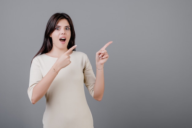 Hermosa mujer morena emocionada y sorprendida señalando copyspace aislado sobre gris