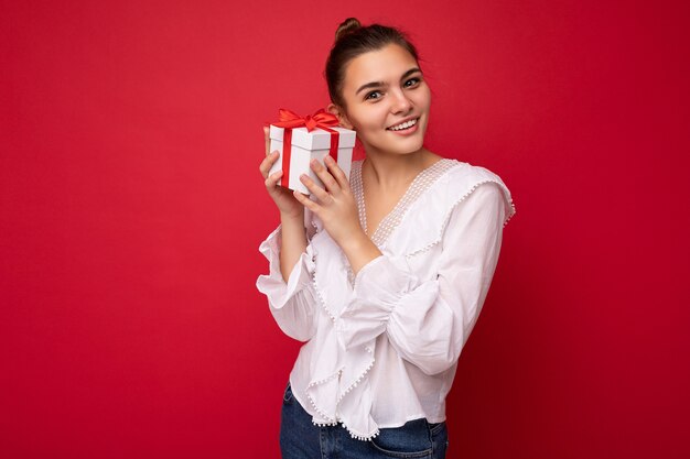 Hermosa mujer morena joven feliz aislada sobre la pared de colores de fondo vistiendo ropa casual elegante con caja de regalo y mirando a la cámara